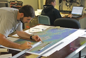 Matt uses four straight edges and a graduated adjustable ruler to plot the ship's course on a large map from latitude and longitude measurements.